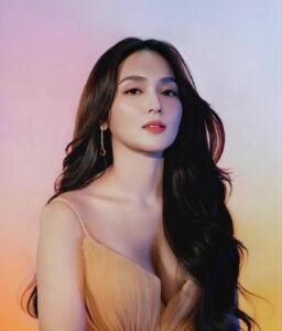 A poised Kathryn Bernardo poses with long dark hair, coral lipstick, and a soft pink-and-yellow gradient background.