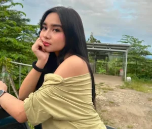 A young woman with long black hair poses outdoors in a light off-shoulder top, resting her chin on her hand near a dirt path.
